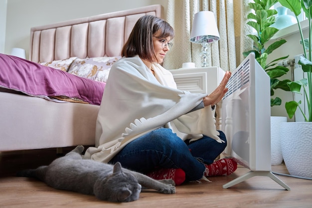 Femme assise sur le sol avec un chat sous une couverture chaude se réchauffant près d'un radiateur électrique