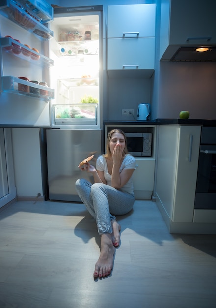 Femme assise sur le sol de la cuisine à côté d'un réfrigérateur ouvert et mangeant une pizza