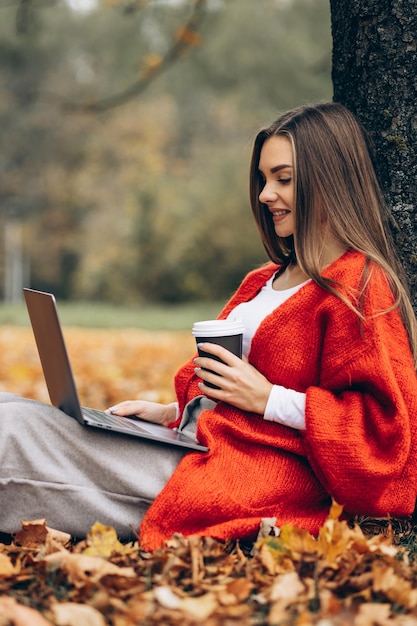 Femme assise sous l'arbre dans le parc travaillant sur un ordinateur portable et buvant du café