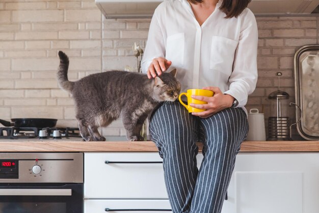 Femme assise sur une table de cuisine avec un chat buvant du thé dans une tasse jaune