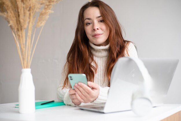 Photo femme assise à table tenant un smartphone en regardant la caméra tout en travaillant à distance ou en apprenant des messages texte