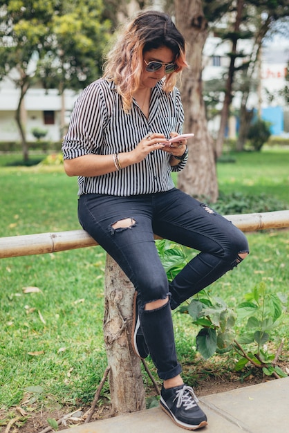 Une femme assise tape sur son smartphone et porte des lunettes de soleil, un haut de pantalon noir à rayures et des baskets noires dans un parc. Image verticale