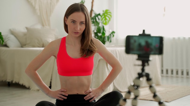 Femme assise sur un tapis et enregistrement vidéo par smartphone sur trépied et faire du yoga à la maison et étirement du corps athlétique