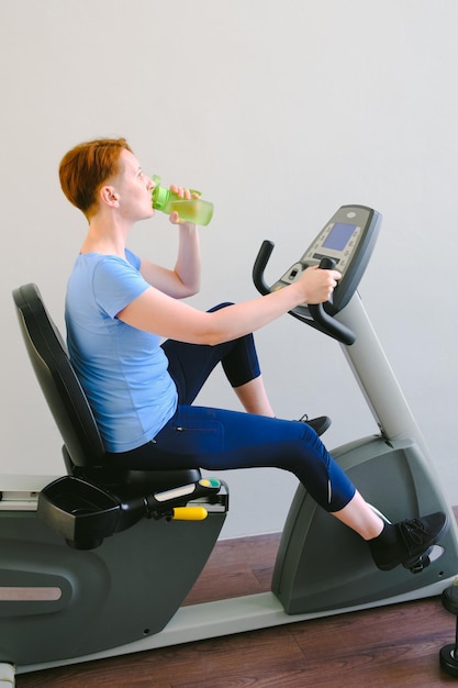 Femme assise sur un vélo d'exercice dans la salle de sport et buvant de l'eau d'une bouteille de sport