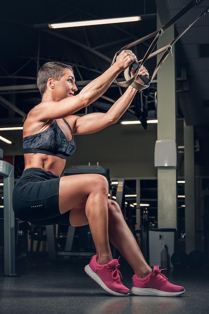 Femme athlétique d'âge moyen aux cheveux courts faisant des entraînements avec des bandes de suspension trx dans un club de gym.