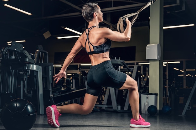 Femme Athlétique D'âge Moyen Aux Cheveux Courts Faisant Des Exercices De Jambes Avec Des Bandes De Suspension Trx Dans Un Club De Gym.