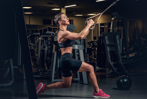 Femme athlétique d'âge moyen aux cheveux courts faisant des exercices de jambes avec des bandes de suspension trx dans un club de gym.