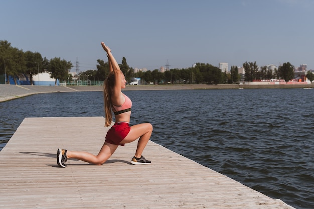 Femme athlétique blonde aux cheveux longs faisant des étirements avant de faire du jogging au bord d'un beau lac