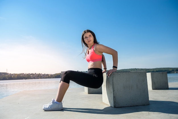 Femme athlétique faisant des exercices accroupis dans un parc ensoleillé près du lac. Concept de bien-être et mode de vie sain