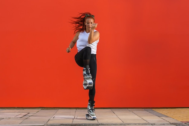 Femme athlétique faisant une routine de remise en forme portant des bottes Kangoo Jumps sur fond orange. Concept de vie saine, d'activité et de sport