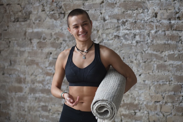 Photo femme athlétique souriante avec tapis d'exercice de yoga debout près du mur de briques, regardant la caméra