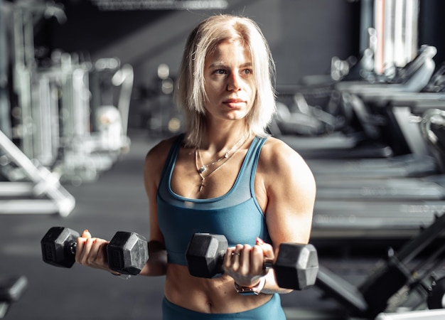 Femme athlétique en tenue de sport forme des biceps avec des haltères lourds dans ses mains au gymnase Concept de fitness et de musculation mode de vie sain