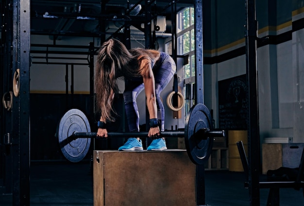 Une femme athlétique vêtue de vêtements de sport colorés se dresse sur une boîte en bois et tient une barre dans un club de gym.