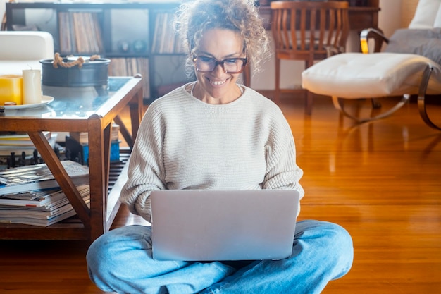 Une femme attrayante assise sur le sol à la maison sourit, regarde l'écran de son portable et parle sur le chat vidéo. Une femme heureuse écoute et parle à un ami.