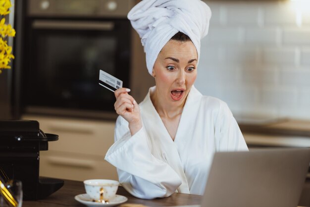 Une femme attrayante en peignoir fait des achats en ligne sur son ordinateur portable tout en dégustant son café du matin et en se relaxant à la maison.