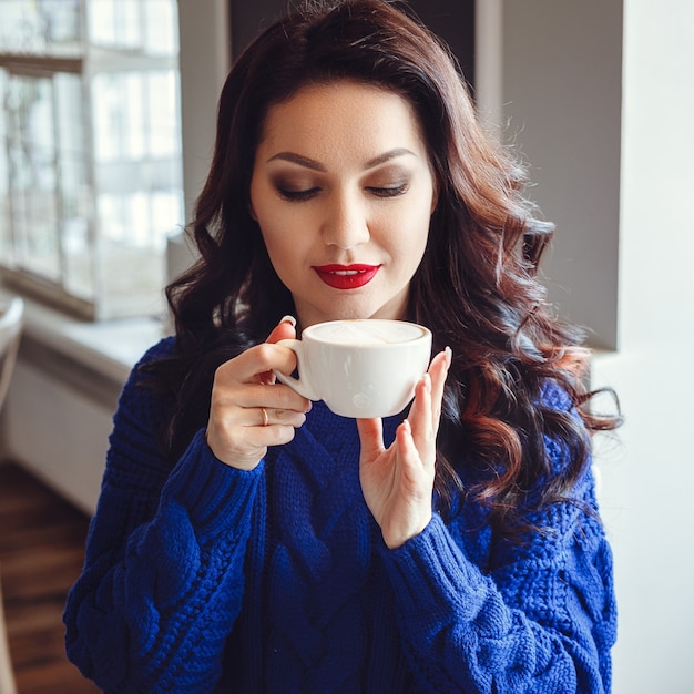 Une femme au café est assise à une table, boit du café et regarde la fenêtre. Femme avec rouge à lèvres et dans un pull bleu attend une réunion, parle au téléphone et sourit beaucoup.