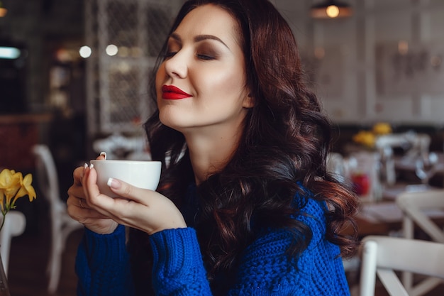 Une femme au café est assise à une table, boit du café et regarde la fenêtre. Femme avec rouge à lèvres et dans un pull bleu attend une réunion, parle au téléphone et sourit beaucoup.