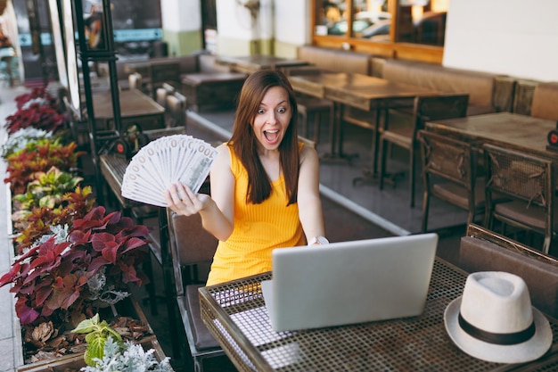 Femme au café de la rue en plein air assis avec un ordinateur portable moderne, tient dans la main un tas de billets de banque en dollars