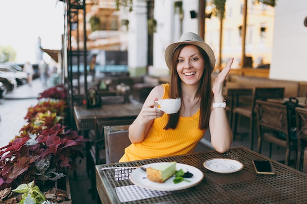 Femme au café de la rue en plein air assis à table dans un chapeau jaune avec une tasse de cappuccino, un gâteau, se détendre au restaurant pendant le temps libre