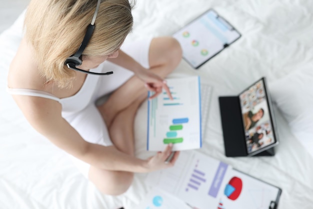 Femme au casque avec des documents dans les mains assise sur le lit et discutant par appel vidéo avec co...