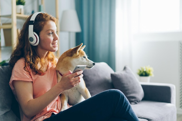 Femme au casque écoutant de la musique caresser un chien sur un canapé tenant un smartphone