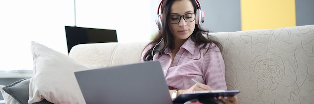 Femme au casque est assise sur un canapé avec un ordinateur portable et tient le presse-papiers dans ses mains