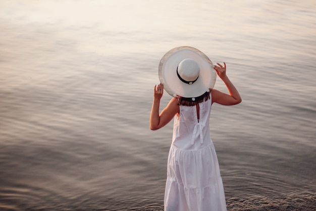 Une femme au chapeau au bord de la mer vue arrière lifestyle portrait d'une femme au chapeau de paille et robe blanche...