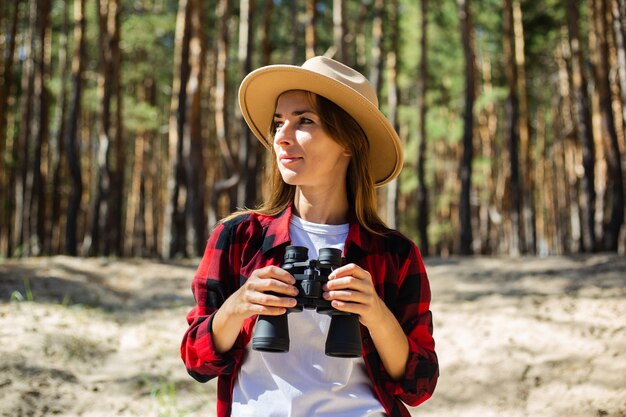 Femme au chapeau et chemise à carreaux rouge tenant des jumelles sur fond de forêt.