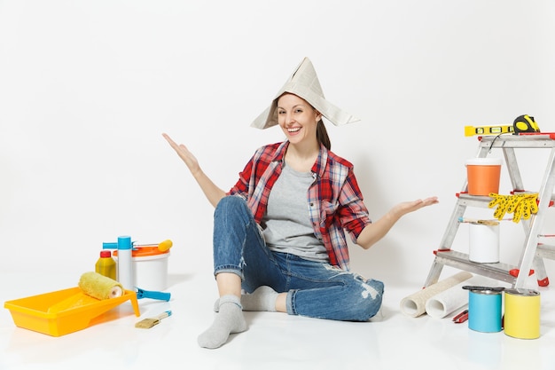 Femme au chapeau de journal écartant les mains, assise sur le sol avec des instruments pour l'appartement de rénovation isolé sur fond blanc. Papier peint, accessoires pour le collage, outils de peinture. Réparer le concept de maison.