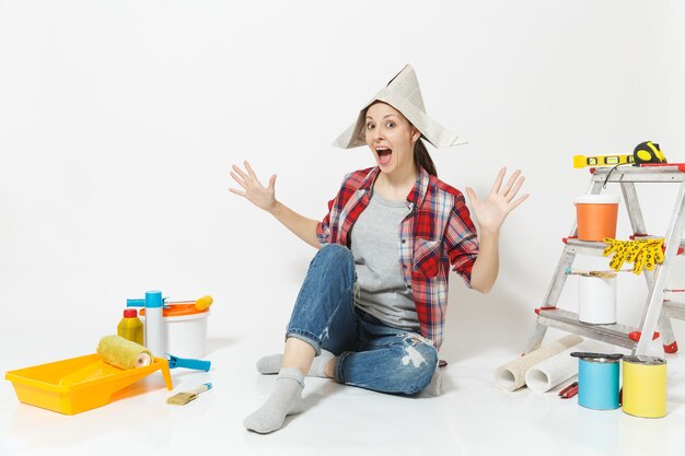 Femme au chapeau de journal écartant les mains, assise sur le sol avec des instruments pour l'appartement de rénovation isolé sur fond blanc. Papier peint, accessoires pour le collage, outils de peinture. Réparer le concept de maison.