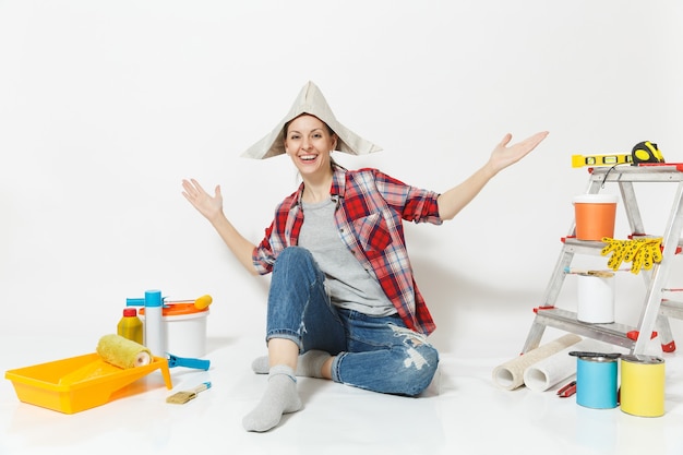 Femme au chapeau de journal écartant les mains, assise sur le sol avec des instruments pour l'appartement de rénovation isolé sur fond blanc. Papier peint, accessoires pour le collage, outils de peinture. Réparer le concept de maison.