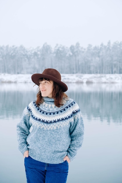 Une femme au chapeau marron et un pull se dresse sur les rives d'un lac et d'une forêt enneigés