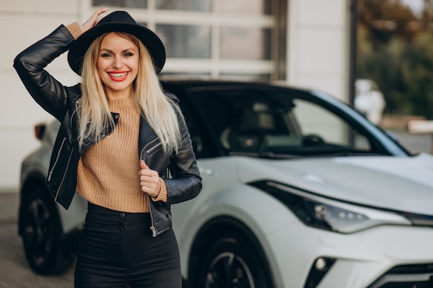 Femme au chapeau noir debout près de sa nouvelle voiture blanche