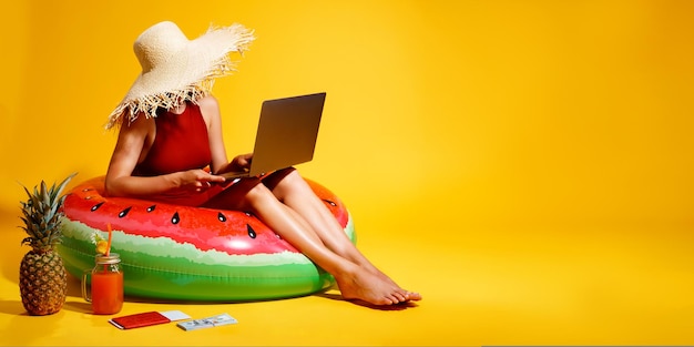 Photo femme au chapeau de paille est assise dans un anneau de pastèque gonflable utilise un ordinateur sur fond jaune concept de voyage