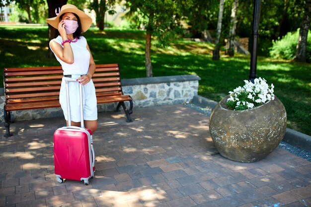 Femme au chapeau de paille et masque de protection rose, dans le parc en plein air avec une valise, parler sur un mobile, la vie pendant la pandémie de coronavirus, ouverture du voyage aérien, concept de voyage.