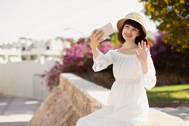 Femme au chapeau et robe blanche utilisant un téléphone intelligent ou prenant un selfie sur la ville tropicale en été