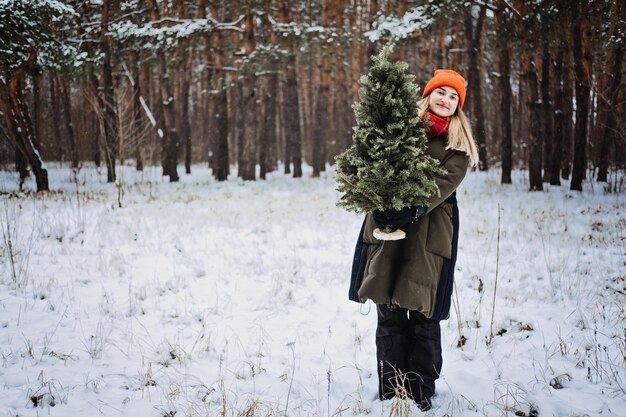 Femme au chapeau rouge tenant un arbre de Noël dans un parc d'hiver de neige se préparant à la cueillette de Noël