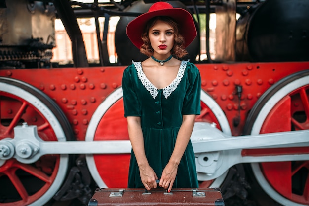 Femme au chapeau rouge avec valise en mains contre locomotive à vapeur vintage, gros plan de roues rouges. Vieux train. Moteur ferroviaire, voyage en train