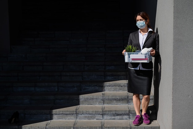 Une femme au chômage portant un masque de protection se tient debout avec une boîte d'effets personnels dans les escaliers Une femme d'affaires licenciée La crise économique mondiale Réduction du travail en raison de la faillite de l'entreprise