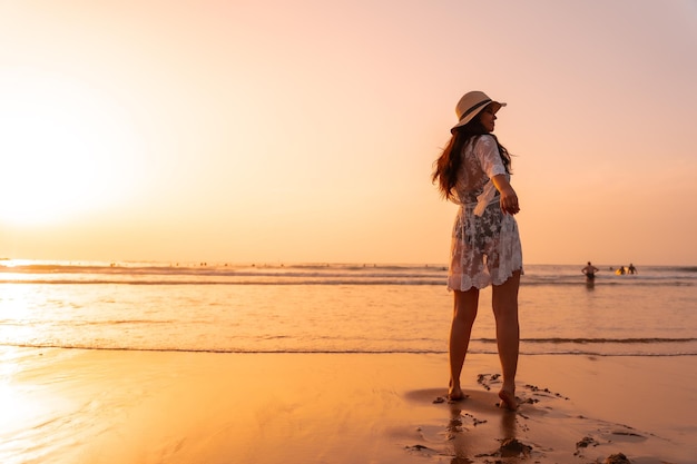 Une femme au coucher du soleil dans une robe blanche à la mer en été avec un chapeau