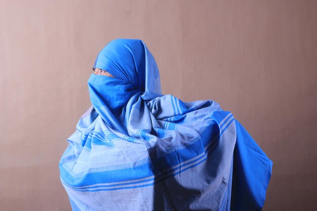 Une femme au foulard bleu se tient devant un mur beige.