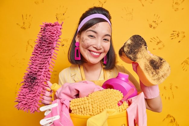 Une femme au foyer asiatique satisfaite porte un bandeau et des boucles d'oreilles a le visage sale après avoir enlevé les poses de poussière avec une éponge à vadrouille nettoie tout dans la maison isolé sur un mur jaune