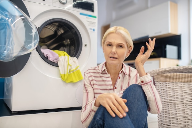 Femme au foyer blonde en chemise rayée assis près de la machine à laver