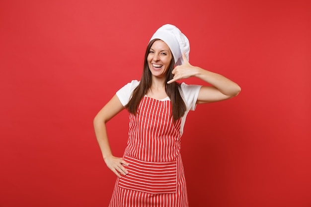 Femme au foyer chef cuisinier ou boulanger en tablier rayé, t-shirt blanc, chapeau de chef toque isolé sur fond de mur rouge. Une femme amusante faisant un geste téléphonique comme dit de me rappeler. Maquette du concept d'espace de copie.