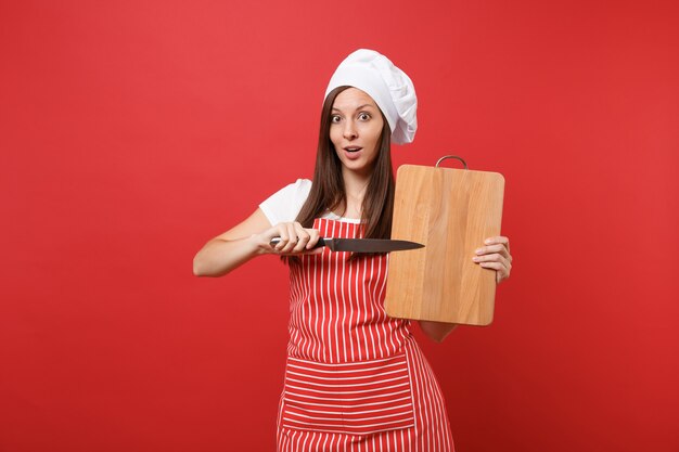Femme au foyer chef cuisinier ou boulanger en tablier rayé, t-shirt blanc, chapeau de chef toque isolé sur fond de mur rouge. Femme de ménage tenant une planche à découper en bois, un couteau. Maquette du concept d'espace de copie.