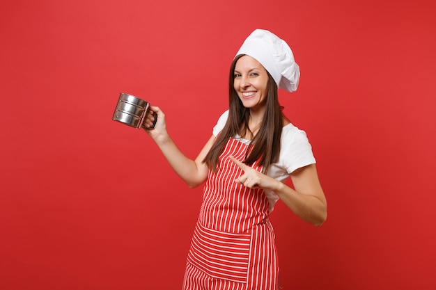 Femme au foyer chef cuisinier ou boulanger en tablier rayé, t-shirt blanc, chapeau de chef toque isolé sur fond de mur rouge. Une femme de ménage tient un tamis en fer métallique, tamisant la farine. Maquette concept d'espace de copie