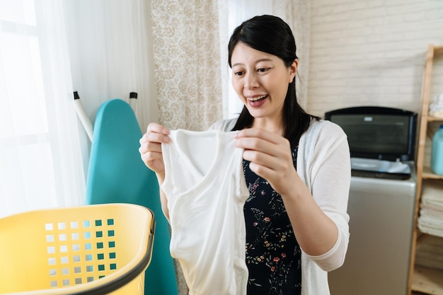 femme au foyer enceinte heureuse asiatique dans la buanderie avec machine à laver. souriante joyeuse future maman en riant tout en regardant des vêtements propres. maternité joyeuse vérifiant les vêtements de bébé dans une maison lumineuse