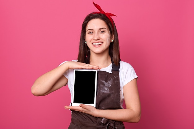 Femme au foyer énergique positive avec de la farine sur le visage et le tablier, tenant la tablette à deux mains