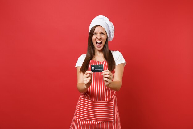 Femme au foyer femme chef cuisinier boulanger en tablier rayé blanc t-shirt toque chapeau de chefs isolé sur fond de mur rouge. Une femme excitée tient en main une carte bancaire de crédit, de l'argent sans numéraire. Maquette concept d'espace de copie