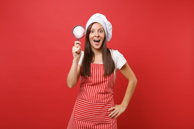 Femme au foyer femme chef cuisinier ou boulanger en tablier rayé t-shirt blanc toque chapeau de chef isolé sur fond de mur rouge. Femme de ménage tenir et regarder à travers une loupe. Maquette concept d'espace de copie